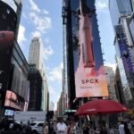 Times Square with SpaceX and Nasdaq billboards, SPCX Nasdaq listed, rocket launch image, people and vehicles on street.