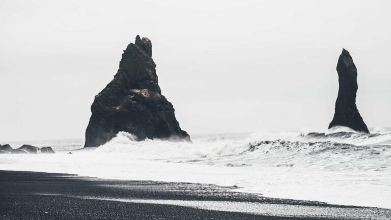 Two large sea stacks rise from the ocean, surrounded by crashing waves and a black sand beach under a cloudy sky.