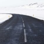Snow-covered landscape with a winding asphalt road and yellow markers, flanked by distant hills under a pale sky.