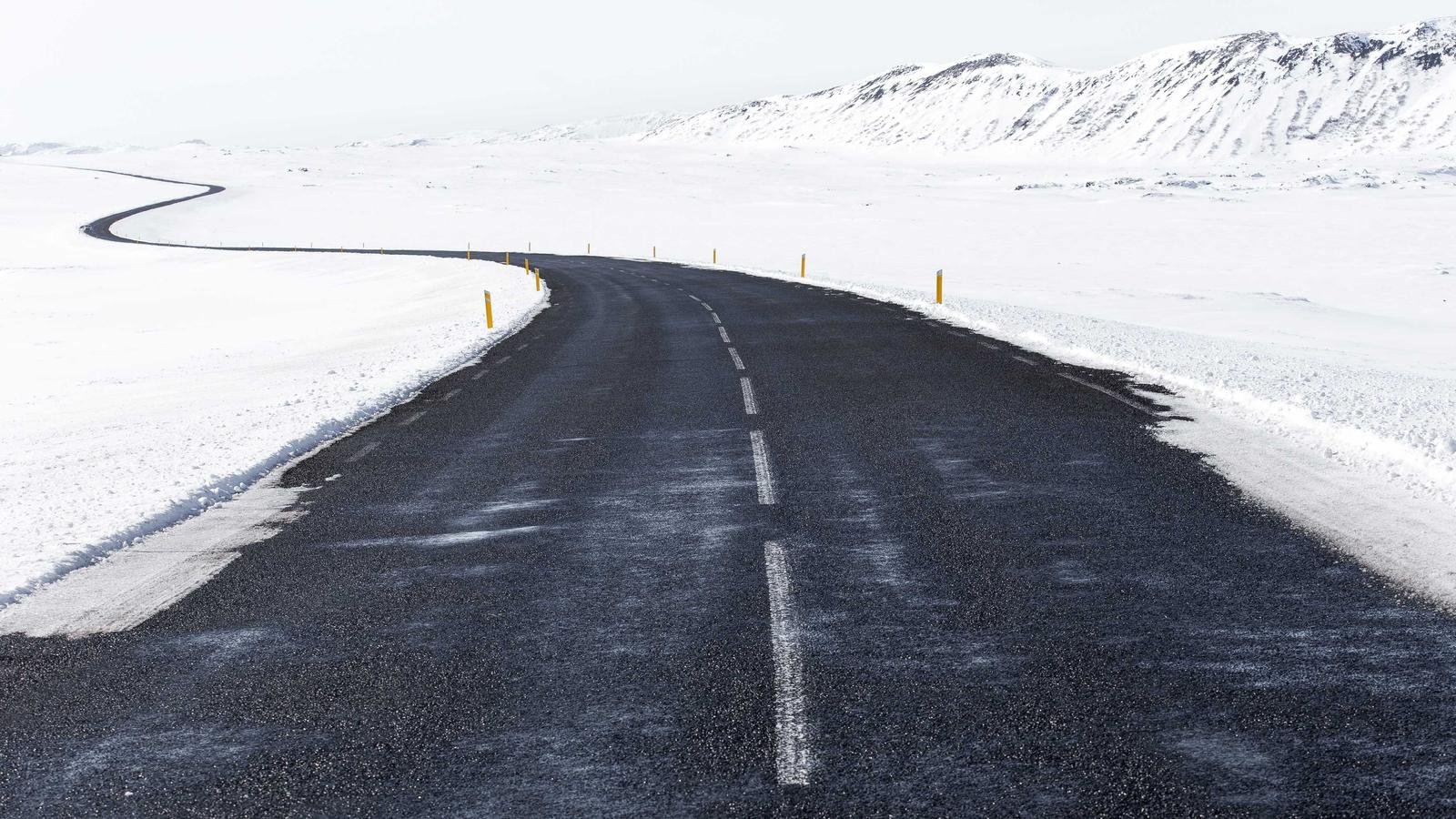 Snow-covered landscape with a winding asphalt road and yellow markers, flanked by distant hills under a pale sky.