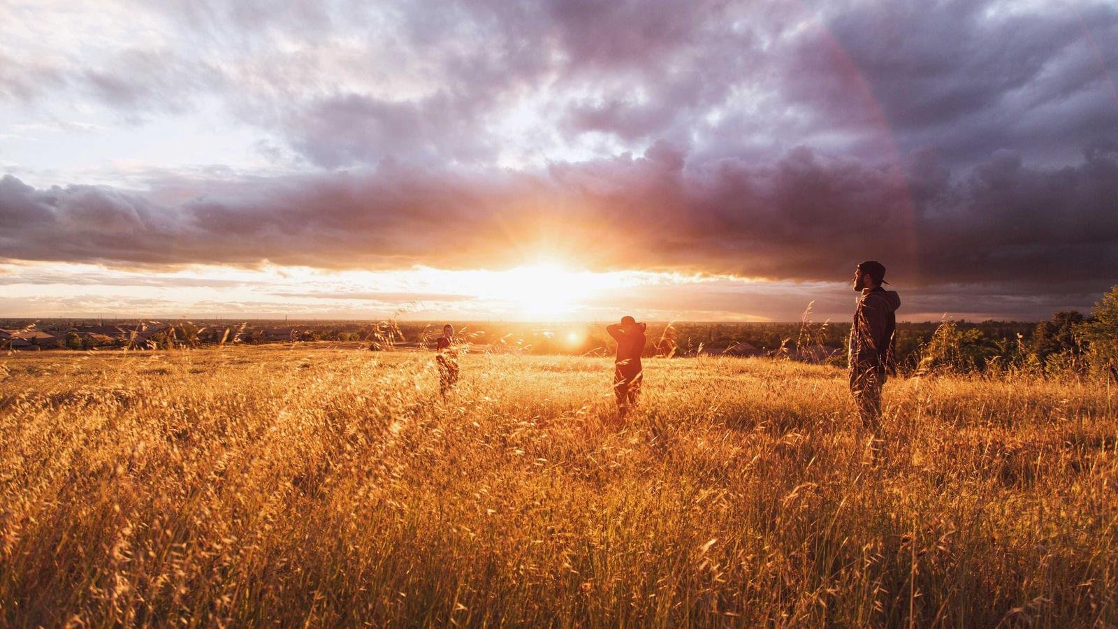 Three people standing in a sunlit field at sunset with dramatic clouds overhead.