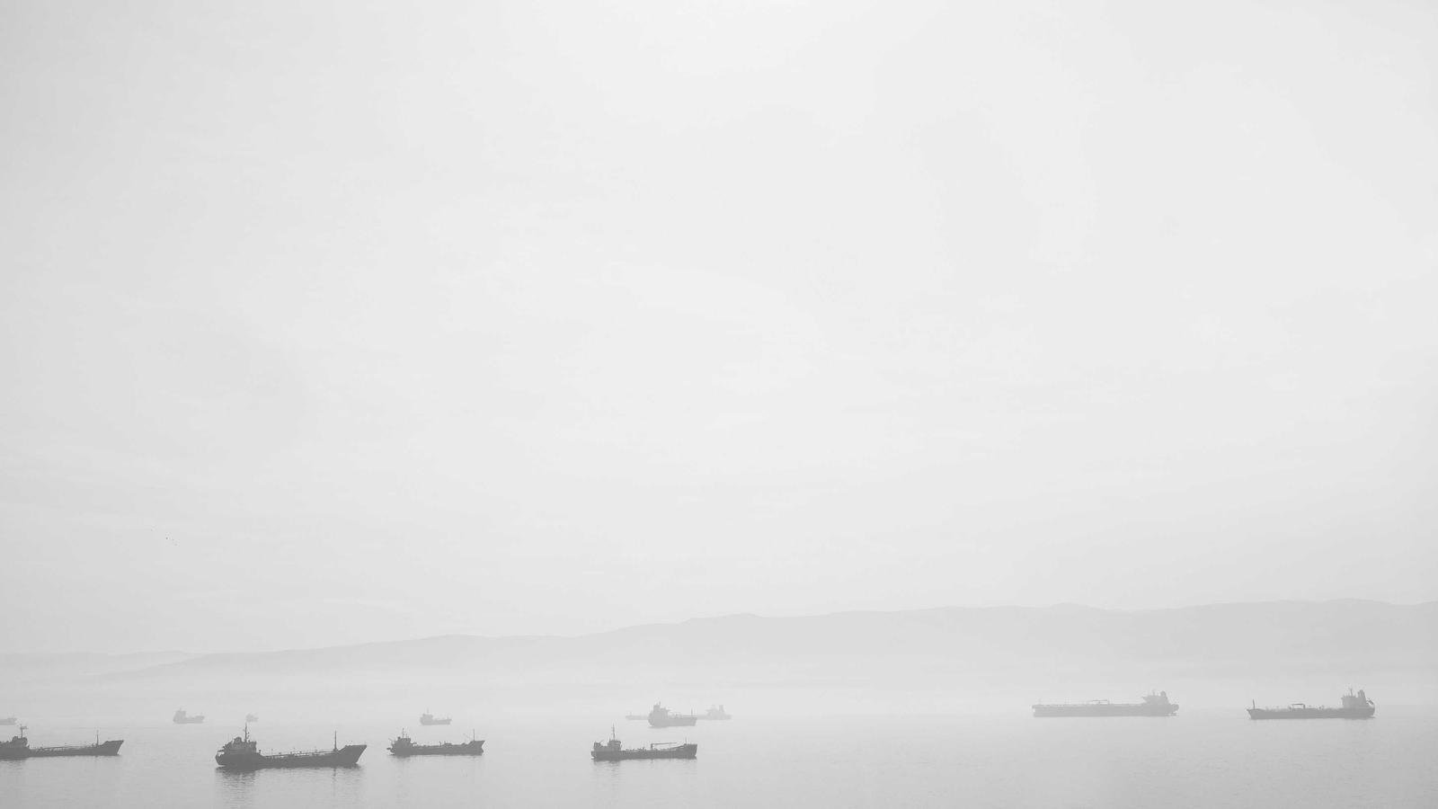 Several ships floating on a foggy sea with distant hills barely visible in the background.