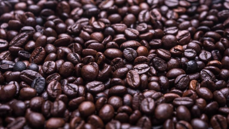 Close-up view of a large pile of roasted coffee beans, showing various shades of brown and glossy textures.