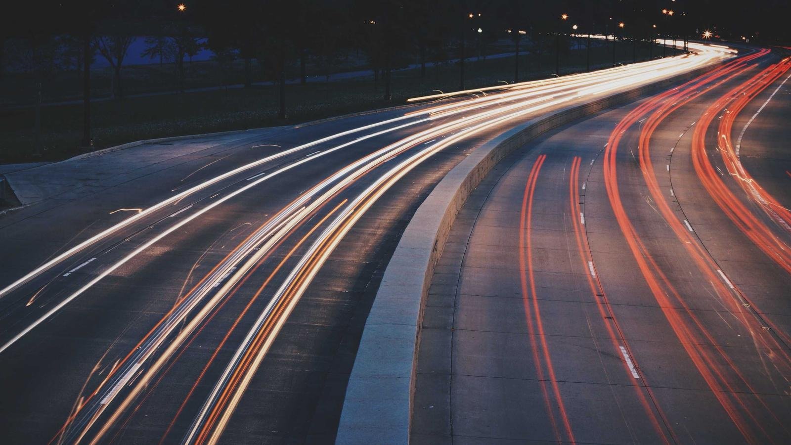 Long exposure of highway at night with red and white light trails showing vehicle motion, streetlights faintly visible.