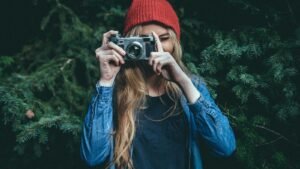 Person in a red beanie and denim shirt taking a photo with a vintage camera, surrounded by green foliage.