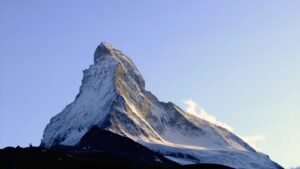 Snow-covered mountain peak under a clear blue sky, with sunlit slopes and a dark foreground.