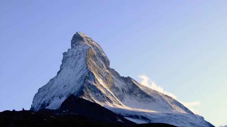 Snow-covered mountain peak under a clear blue sky, with sunlit slopes and a dark foreground.