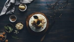 Cake topped with poached pears and leaves on a wooden board, surrounded by bowls of ingredients and a cloth on a dark surf...