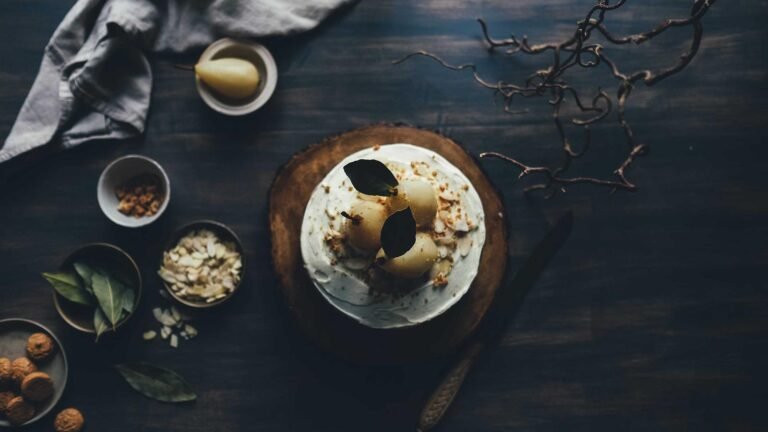 Cake topped with poached pears and leaves on a wooden board, surrounded by bowls of ingredients and a cloth on a dark surf...