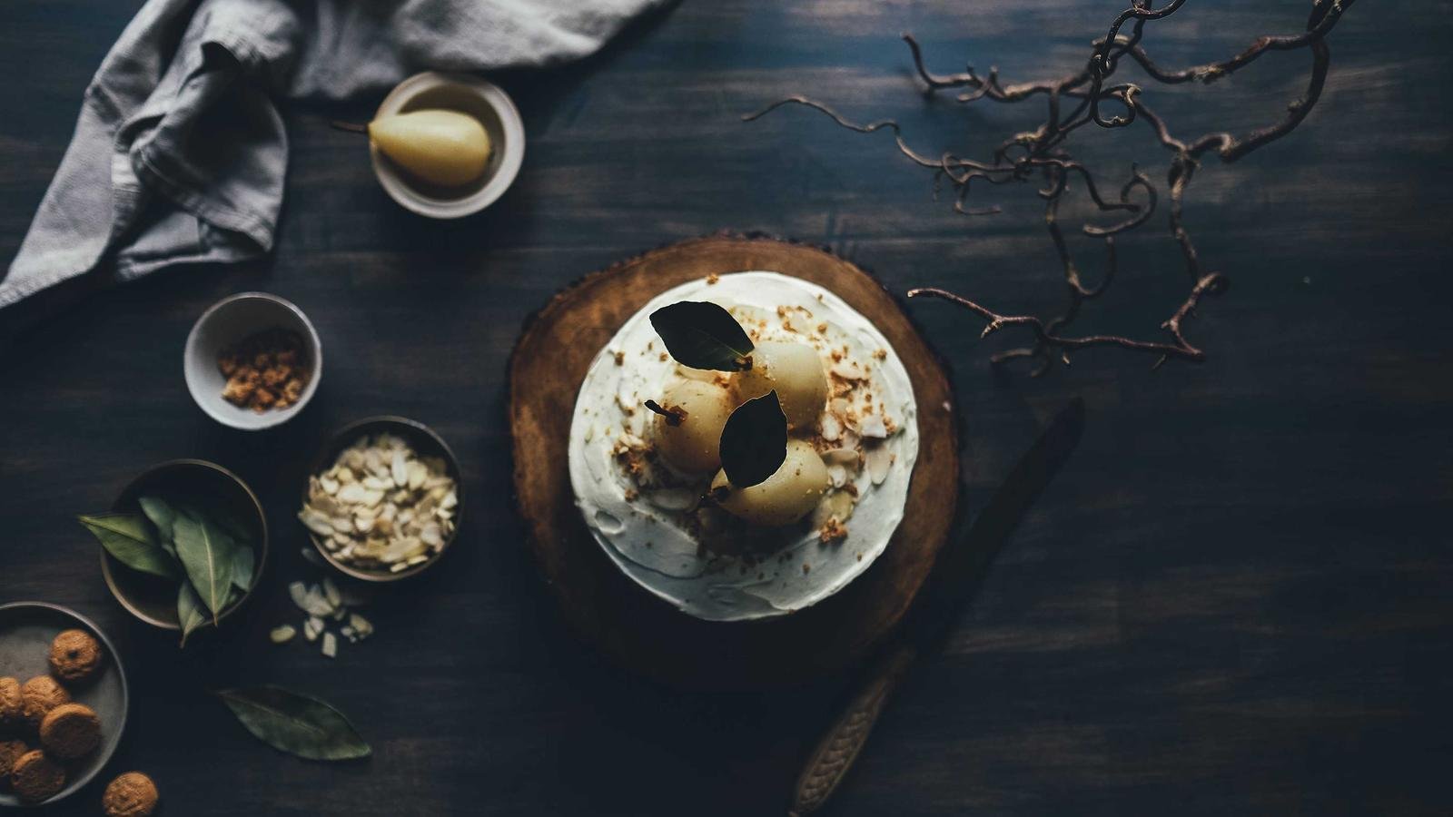 Cake topped with poached pears and leaves on a wooden board, surrounded by bowls of ingredients and a cloth on a dark surf...