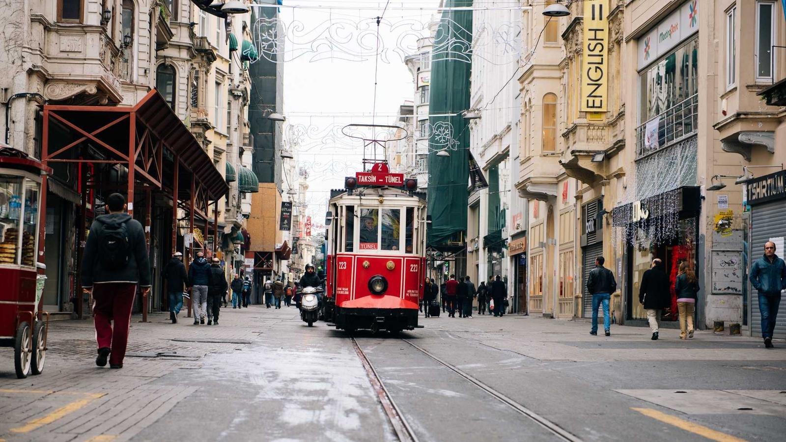 Red tram labeled "Taksim-Tünel" on a busy street with pedestrians and shops, including a sign for English courses.