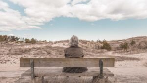 Person with gray hair sitting on a wooden bench, facing a sandy landscape with sparse vegetation under a cloudy sky.