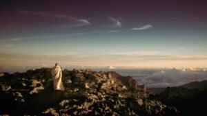 Person wrapped in a blanket stands on rocky terrain overlooking clouds and a sunset sky.