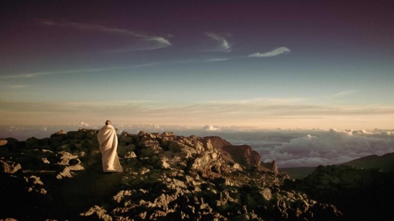 Person wrapped in a blanket stands on rocky terrain overlooking clouds and a sunset sky.