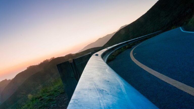 Curved road with metal guardrail winding through hills at sunset, with pink and orange hues in the sky.