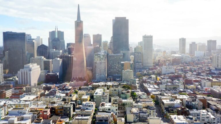 San Francisco skyline with iconic Transamerica Pyramid and surrounding skyscrapers under a bright, partly cloudy sky.
