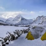 Snow-covered mountains and a yellow tent in a snowy landscape, with a small stone hut nearby under a clear blue sky.