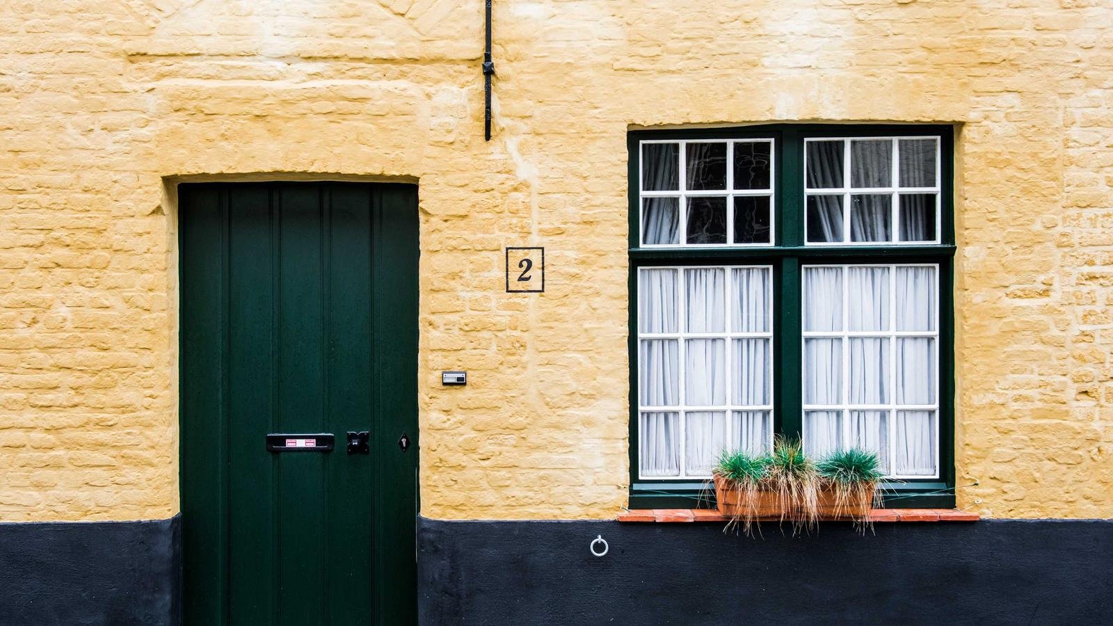 Yellow brick wall with a green door, number 2, a window with white curtains, and a planter with grass.