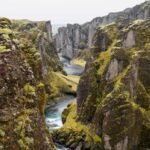 Lush green cliffs with a winding river flowing through a narrow canyon under a cloudy sky.