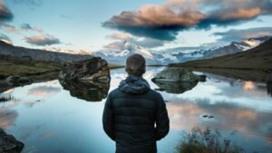 Person in a black jacket stands by a calm lake, surrounded by mountains and a dramatic cloudy sky at sunset.