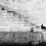 Two people sitting separately on stone steps against a large brick wall, one higher and one lower, in a black and white se...