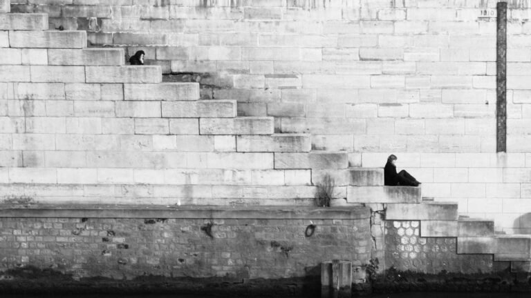 Two people sitting separately on stone steps against a large brick wall, one higher and one lower, in a black and white se...
