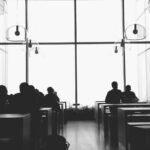 Silhouettes of people seated at tables in a modern, high-ceilinged room with large windows and hanging light fixtures.
