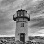 Black and white lighthouse stands on rocky hill under a cloudy sky, surrounded by open landscape.