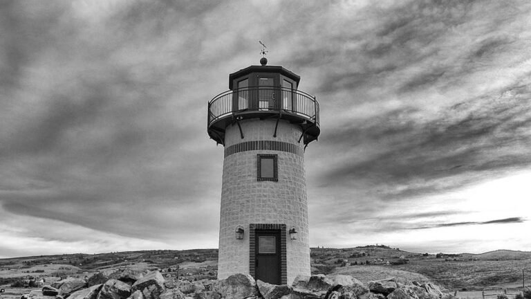 Black and white lighthouse stands on rocky hill under a cloudy sky, surrounded by open landscape.