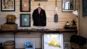 Store display with jacket, shirts, backpack, framed art, paddle, lamp, and plant on wooden shelves against a paneled wall.