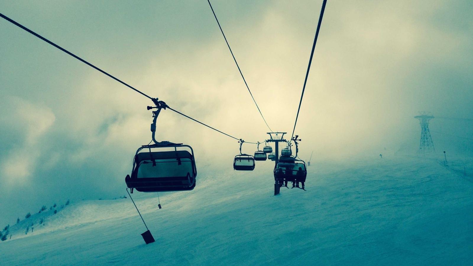 Ski lift chairs ascending a snowy mountain under a cloudy sky, with distant skiers visible on the slope.