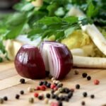 Sliced red onion, peppercorns, parsley, and mixed vegetables on a wooden cutting board.