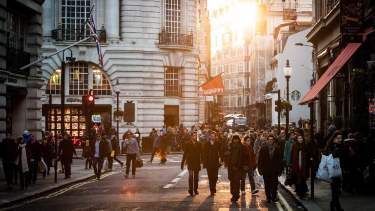 Busy city street with people walking, a red traffic light, and storefronts including Superdry and Starbucks under warm sun...