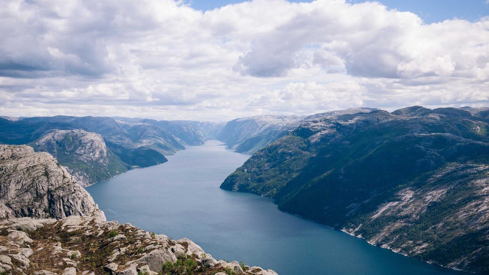 Expansive view of a fjord with steep rocky cliffs and a clear blue sky filled with scattered clouds.