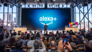 Person on stage presenting Alexa+ in front of large audience, backdrop shows Alexa+ logo on screen, attendees taking photos.