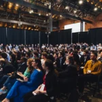 A large audience seated in a conference hall with high ceilings and black curtains, attentively watching a presentation.