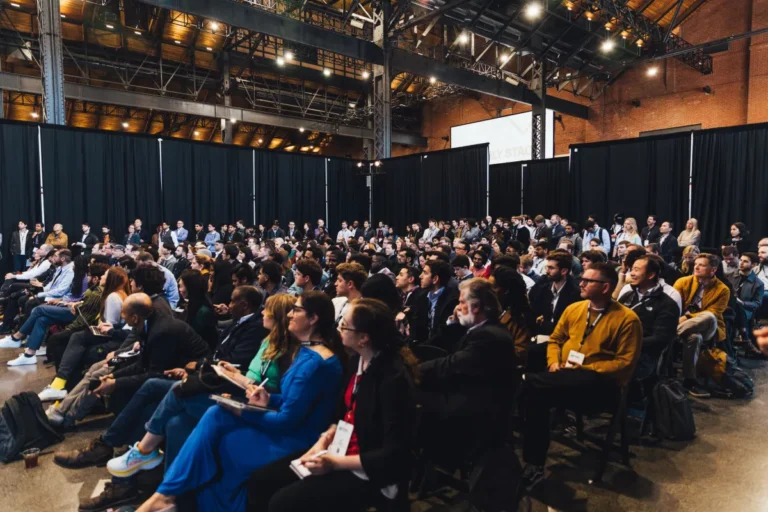 A large audience seated and standing in a spacious industrial venue, attentively watching a presentation, with black curta...