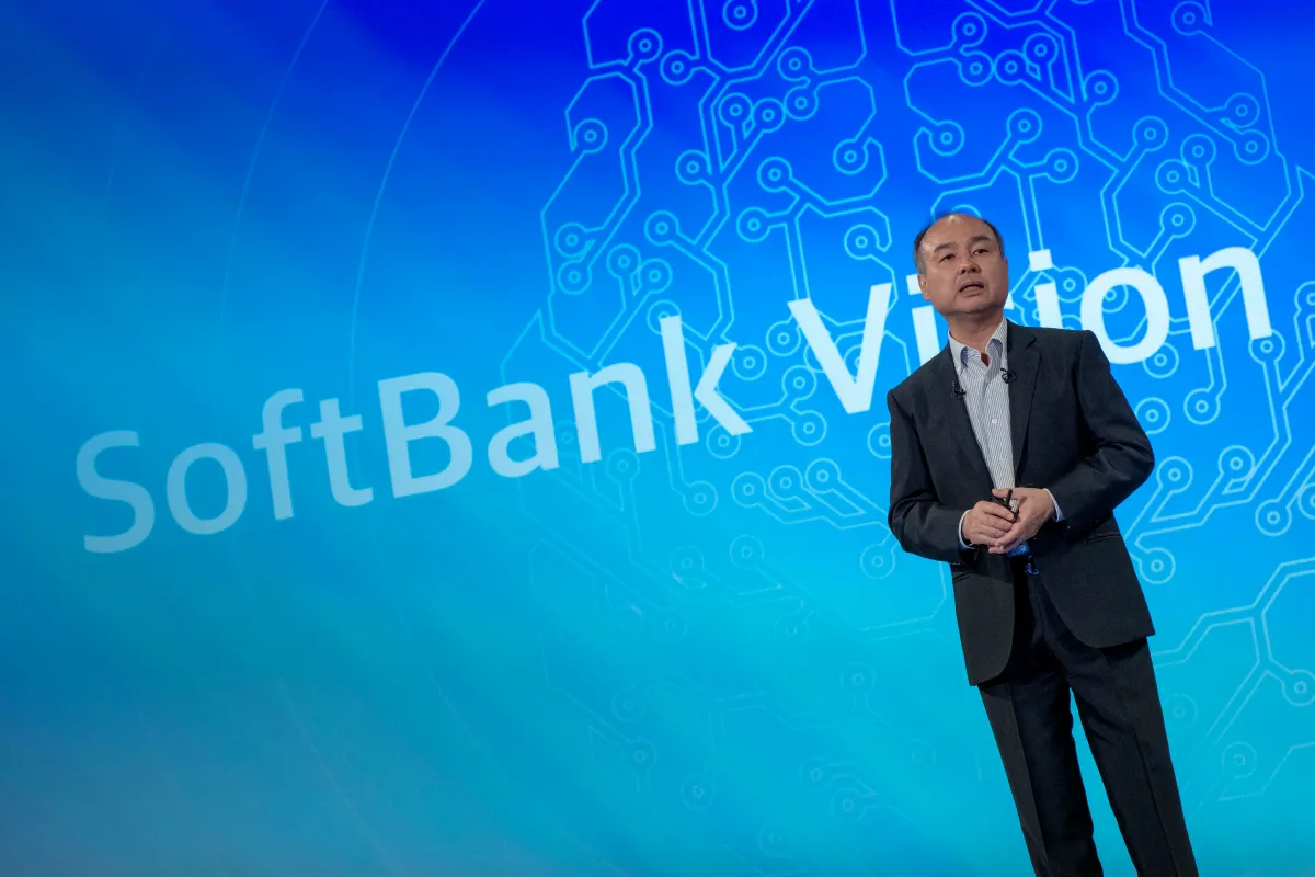 Man in a suit stands on stage with a blue circuit pattern background and "SoftBank Vision" text.