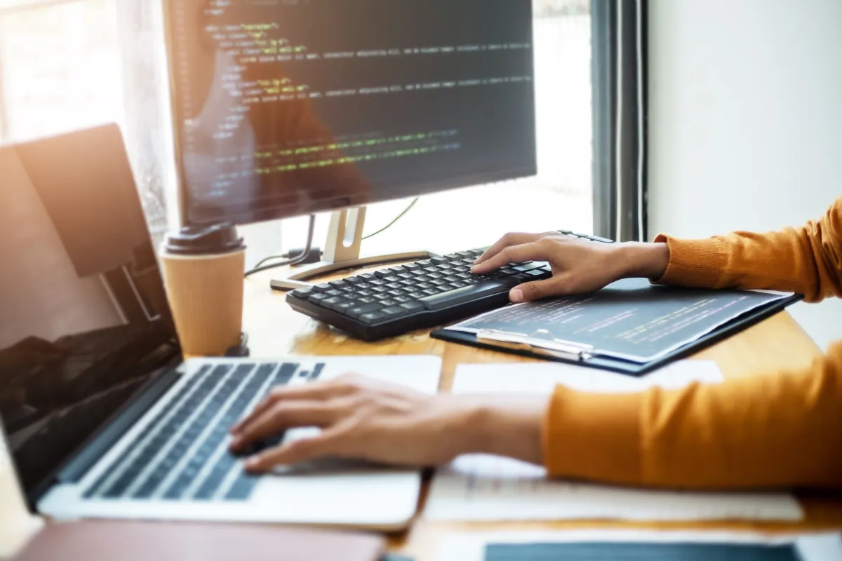 Person typing on a laptop with code on a monitor, coffee cup nearby, in a bright office setting.