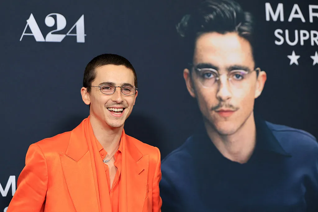Person in an orange suit smiles beside a large poster of another person. A24 logo visible in the background.