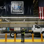 Worker in a factory beside a white truck chassis, large "Built Ford Tough" banner and American flag in the background.