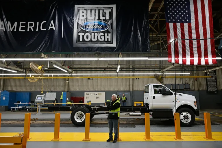 Worker in a factory beside a white truck chassis, large "Built Ford Tough" banner and American flag in the background.