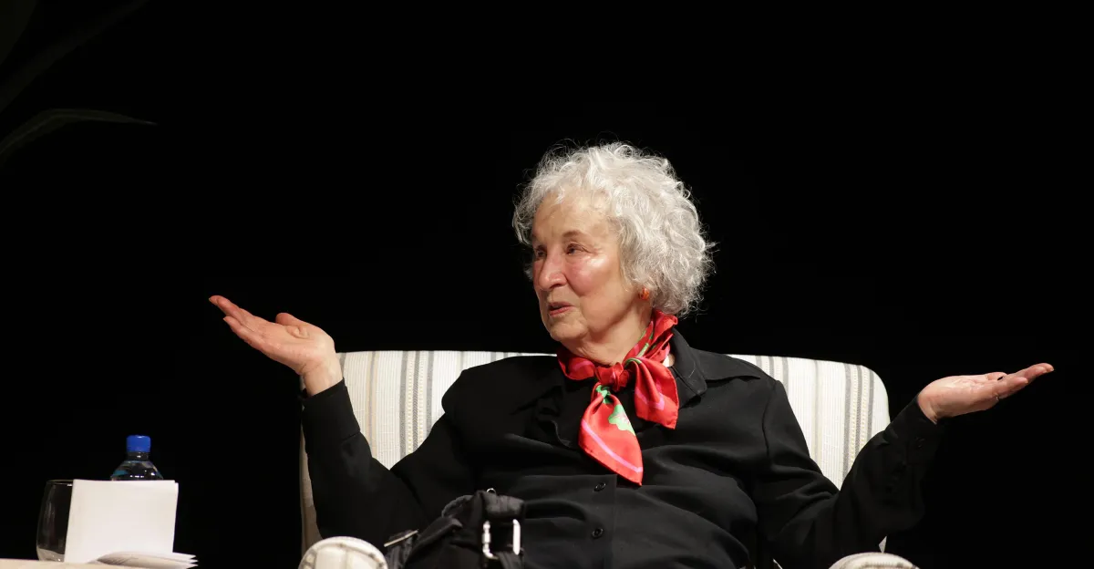 Elderly woman with curly hair gestures with open hands, sitting on a striped chair, wearing a red scarf and black outfit.