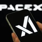 Hand holding a smartphone displaying a white logo on a black screen, blurred spacex logo in the background