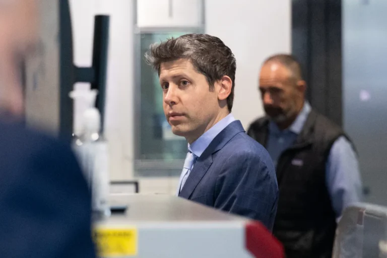 Man in a suit standing indoors, looking off-camera, with another man in the background. Office setting with a monitor.