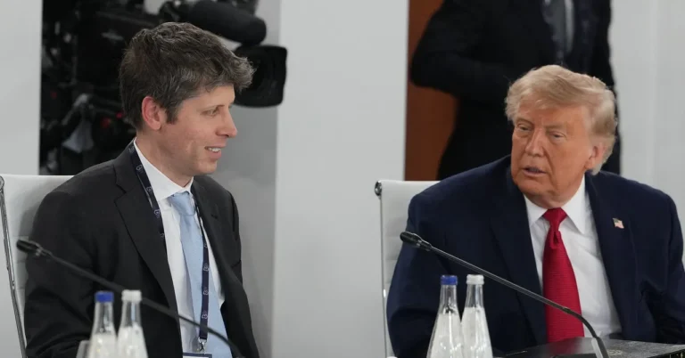 Two men seated at a conference table, engaged in conversation, with cameras and microphones in the background.