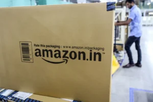 Large Amazon.in box with packaging text in a warehouse; a person in the background handles packages.