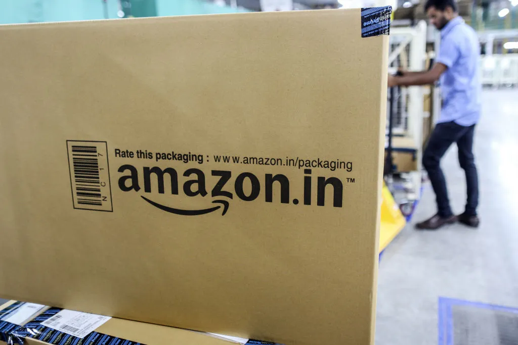 Large Amazon.in box with packaging text in a warehouse; a person in the background handles packages.