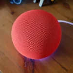 Red spherical smart speaker with mesh design on wooden table, connected by a white cable.