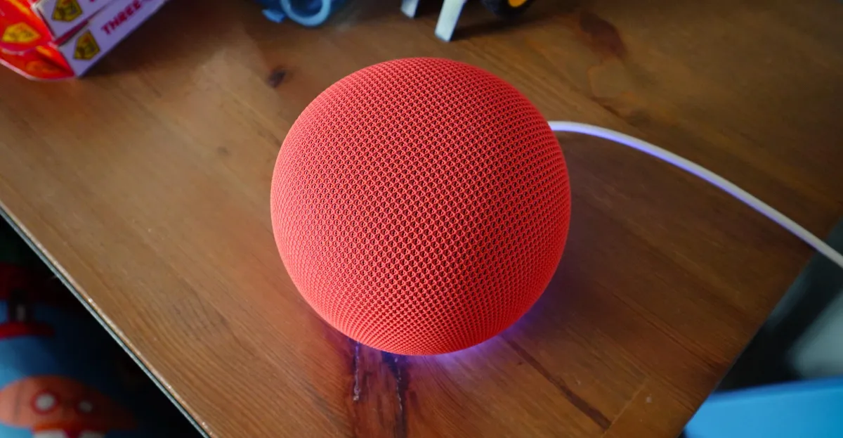 Red spherical smart speaker with mesh design on wooden table, connected by a white cable.